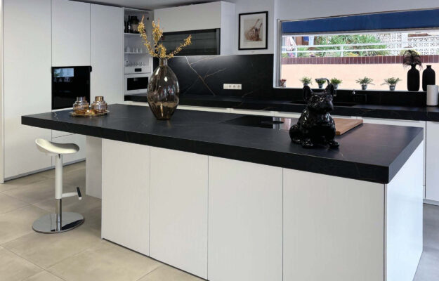 Foto der Ausstellung des Küchenstudios: Weiße L-Küche mit Kochinsel und schwarzer Steinarbeitsplatte.