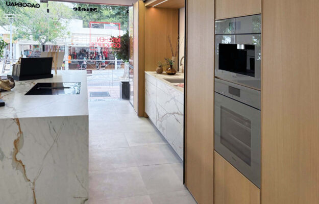 Blick auf eine schwarze Arbeitsplatte im Vordergrund. Im Hintergrund ist eine weiße marmorne Kochinsel von Holzschränken zu sehen.