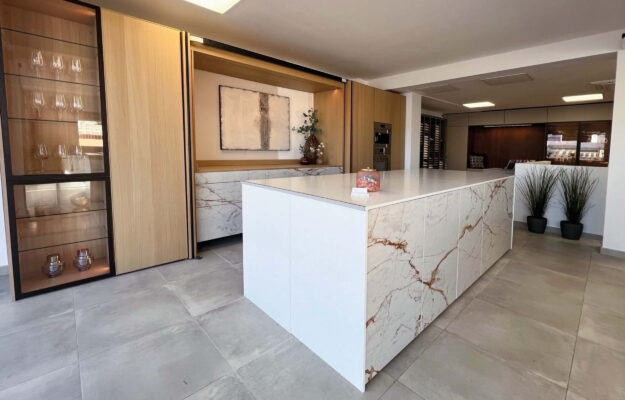 Weiße Kochinsel vor einer Küchenzeile mit Hochschränken aus Holz.